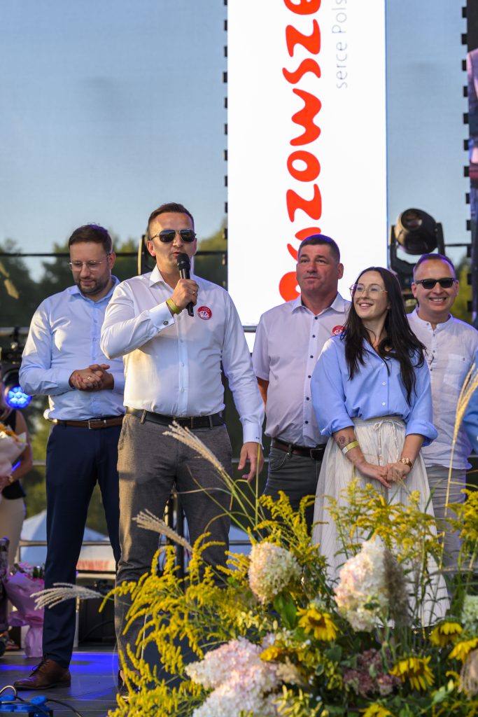 Grupa osób na scenie, jedna osoba mówi do mikrofonu. W tle duży biały ekran z czerwonym napisem. Na przodzie stoją żółte i białe kwiaty.