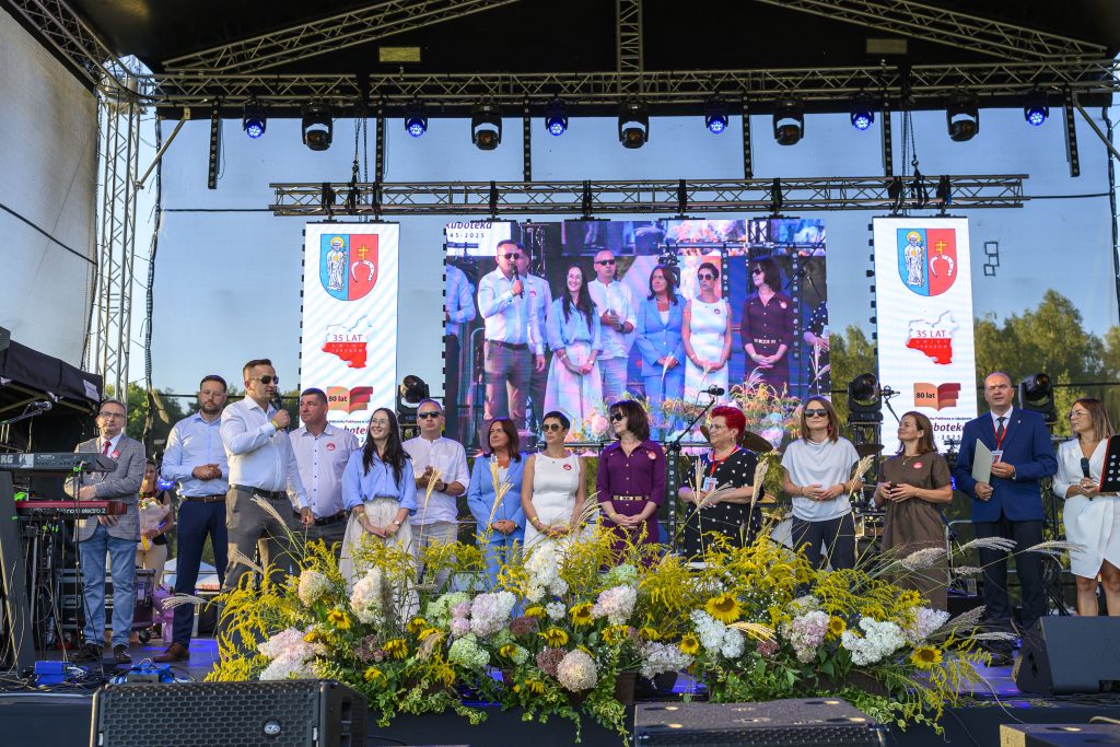 Grupa ludzi na scenie, w tle duży ekran. Kwiaty w przedniej części sceny. Jasne, słoneczne otoczenie.