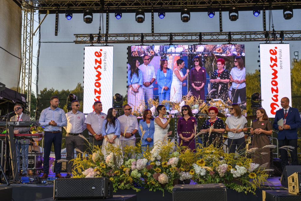 Grupa ludzi stoi na scenie ozdobionej kwiatami, przed dużym ekranem. Ludzie są elegancko ubrani, tło stanowią drzewa. Na scenie znajdują się także dwa banery z napisem Mazowsze, serce Polski.