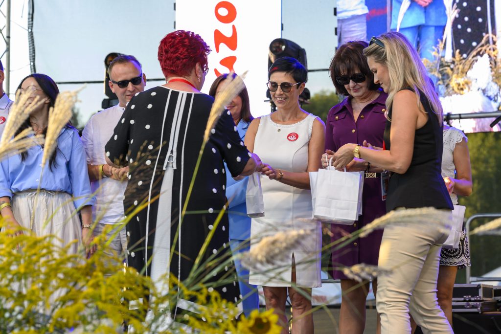 Grupa osób stoi na scenie, każda trzyma białą torbę. Kolejka osób ubrana jest kolorowo, między innymi w białe i fioletowe stroje. Na pierwszym planie widać żółte trawy i zielone rośliny.