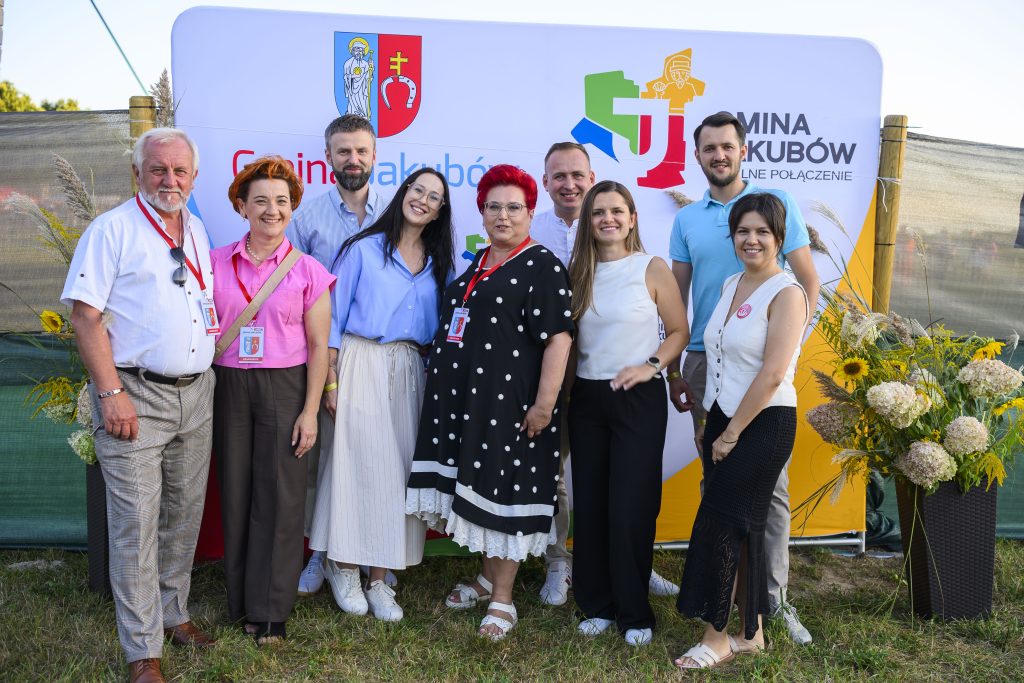 Grupa ludzi stoi na trawie przed banerem gminnym. Tło zdobią kolorowe logo oraz kwiaty. Strój uczestników jest zróżnicowany i kolorowy.