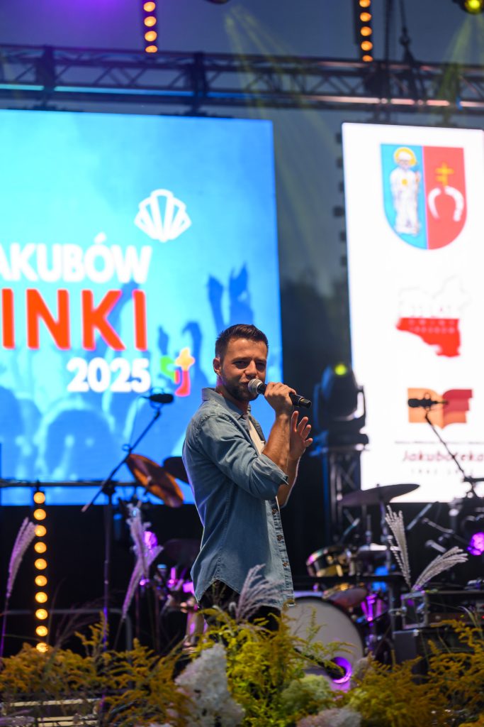 Na scenie mężczyzna śpiewa do mikrofonu w niebieskiej koszuli. W tle widoczne są bębny i jasny ekran z napisem. Przed sceną dekoracja z zieleni i białych kwiatów.