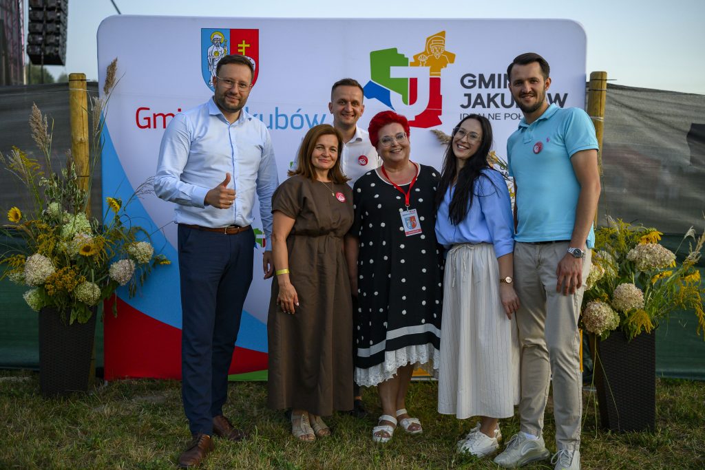 Na zdjęciu grupa sześciu osób stoi przed kolorowym banerem z napisami i logiem gminy Jakubów. Baner jest biały z czerwonymi i zielonymi elementami, otoczony bukietami kwiatów. Ludzie są uśmiechnięci, a tło to zielona