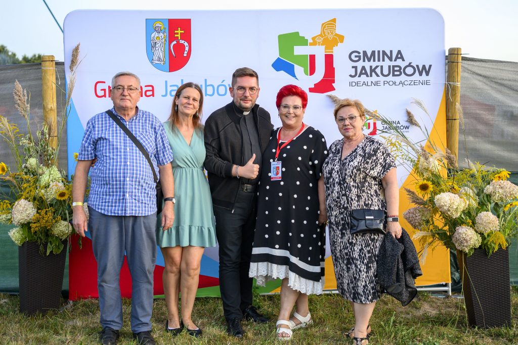Pięć osób stoi na tle kolorowego baneru z napisem Gmina Jakubów. Tło zdobią kwiatowe aranżacje z żółtymi słonecznikami. Osoby ubrane są w letnie stroje.