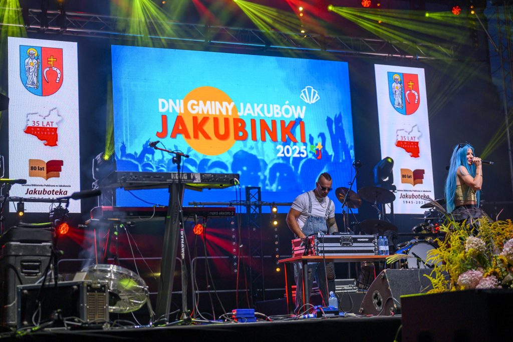 Muzyczny występ na scenie. Kolorowa grafika na ekranie z napisem Dni Gminy Jakubów Jakubinki 2025. Światła i instrumenty w tle.