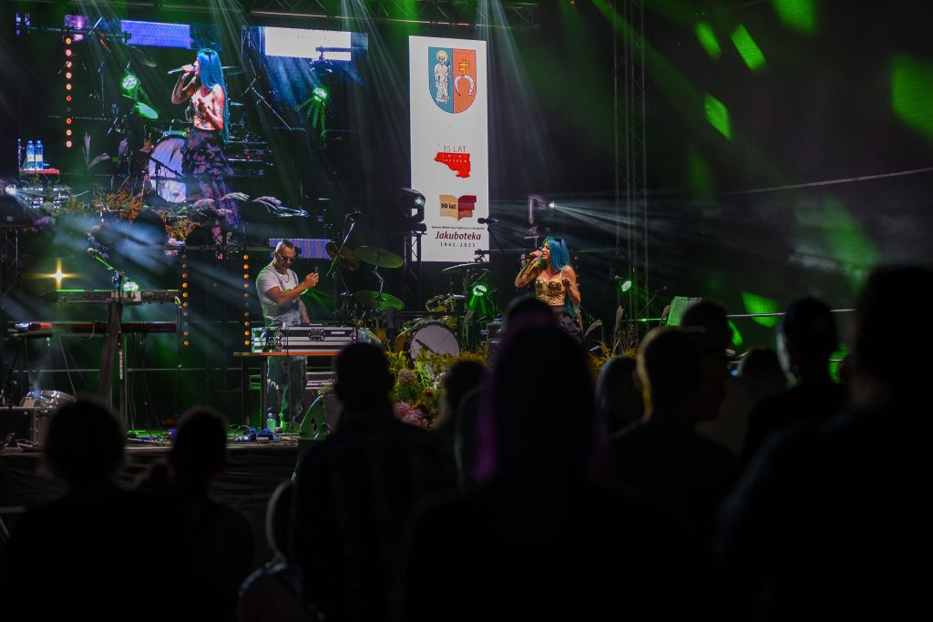 Na scenie odbywa się koncert z kolorowym oświetleniem. Występuje kobieta z niebieskimi włosami i mężczyzna przy sprzęcie DJ. W tle widoczne są bębny i ekran z logo i napisami.