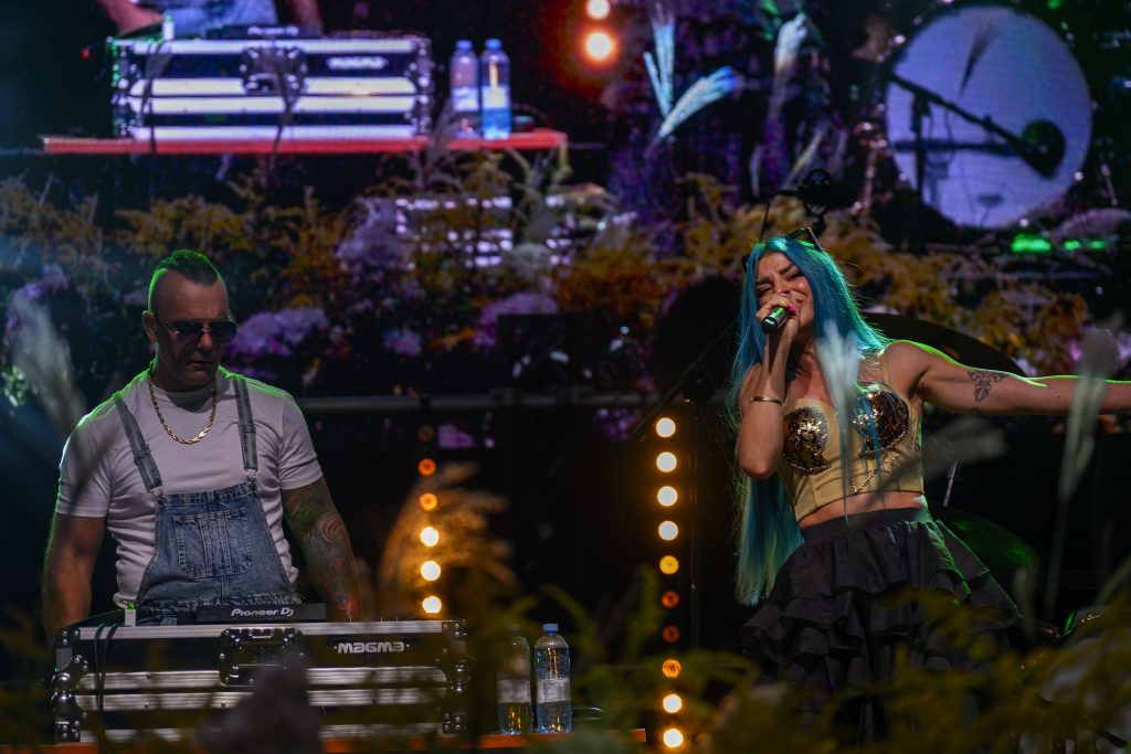 Kobieta z niebieskimi włosami śpiewa na scenie, obok DJ w okularach. Kolorowe światła i tło o tematyce roślinnej.
