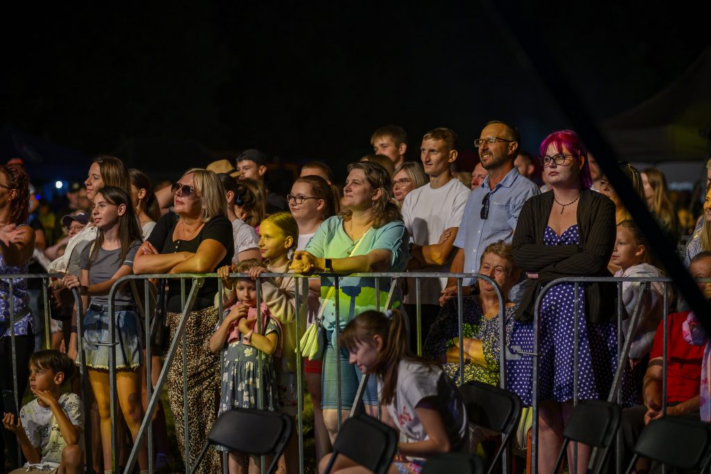 Grupa ludzi na koncercie na świeżym powietrzu, stojąca za metalowym ogrodzeniem. Tło ciemne, scena oświetlona. Uczestnicy ubrani różnorodnie, kobieta z różowymi włosami.