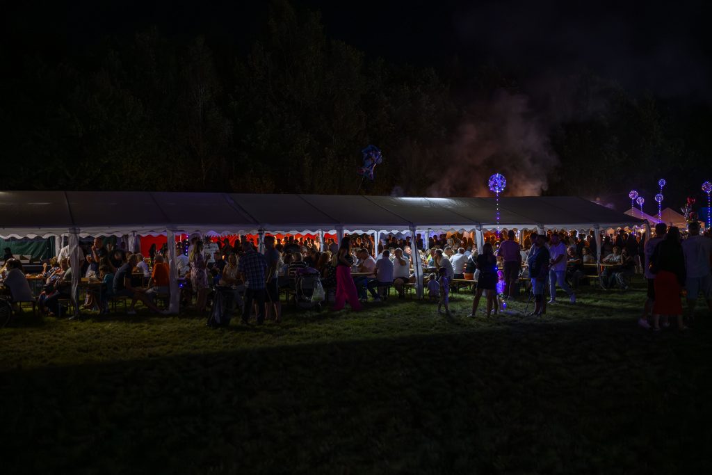 Nocna scena z ludźmi pod długim, białym namiotem, podświetlanym na czerwono. W tle ciemność i dym. Kolorowe światła wokół.