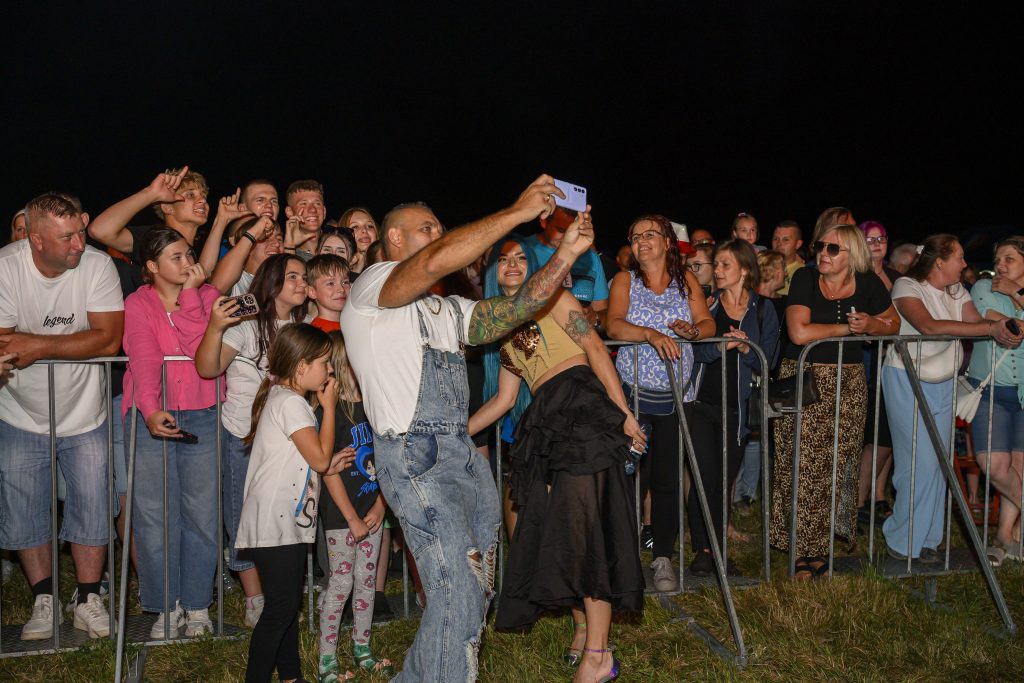 Grupa ludzi robi selfie nocą. Mają kolorowe ubrania i stoją za barierkami. Tło jest ciemne, podkreślając zebranych.