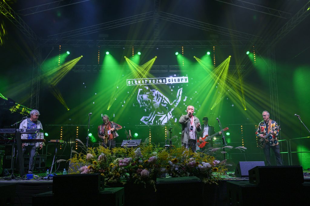 Zespół gra na scenie oświetlonej zielonymi światłami. W tle widoczny jest neonowy napis i ekran z grafiką. Na przodzie sceny są kwiaty.