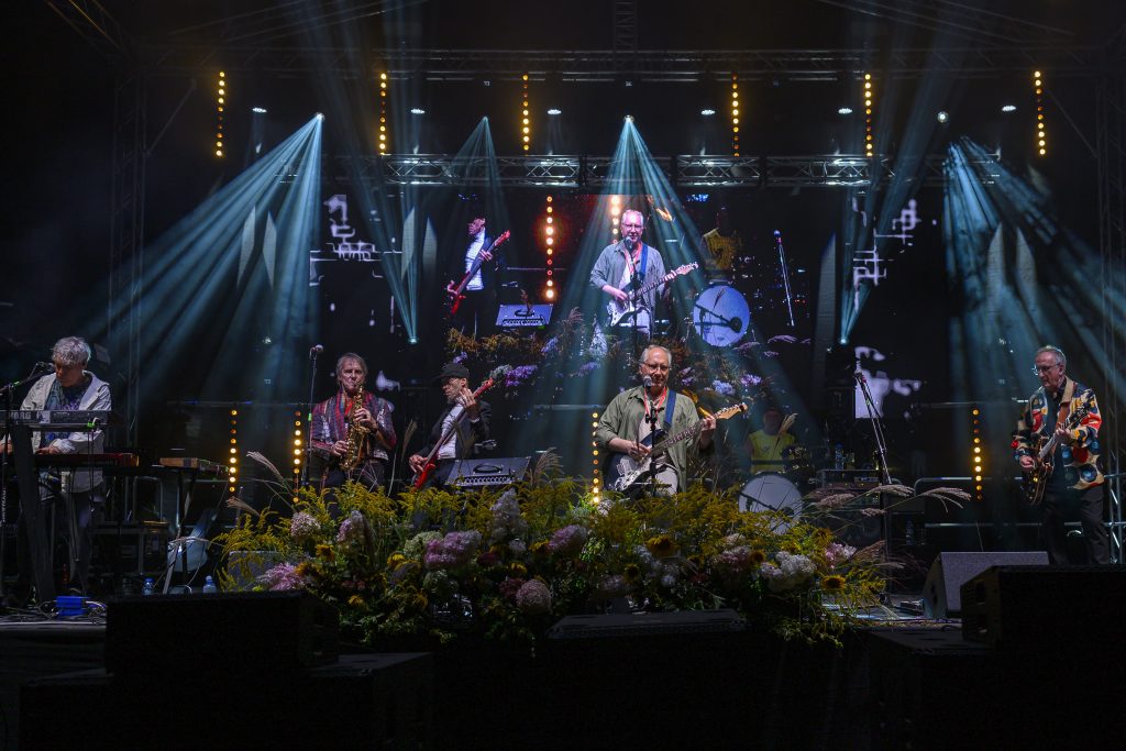 Na zdjęciu zespół na scenie podświetlonej niebieskimi i żółtymi światłami. W tle duży ekran z projekcją muzyków. Przód sceny zdobią kolorowe kwiaty.