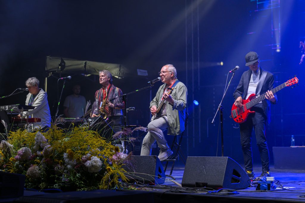 Zespół muzyczny na scenie gra koncert. Artyści otoczeni są kolorowymi światłami i kwiatowymi dekoracjami. Tło sceny jest ciemne.
