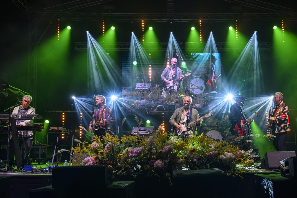 Na scenie występuje zespół muzyczny z instrumentami. W tle zielone światła i ekran z muzykami. Przód sceny zdobią kwiaty.