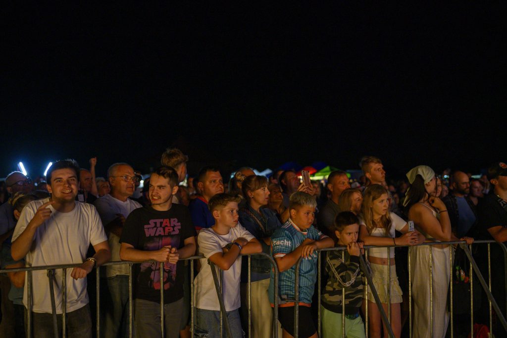 Tłum ludzi na koncercie nocą, oświetlonych ciepłym światłem. Czarny mrok w tle. Różnorodna grupa osób przy barierkach.