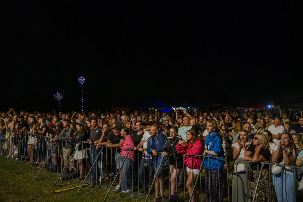Tłum ludzi podczas koncertu na zewnątrz w nocy. Ubrania są różnokolorowe. W tle czarne niebo, kilka kolorowych świateł.