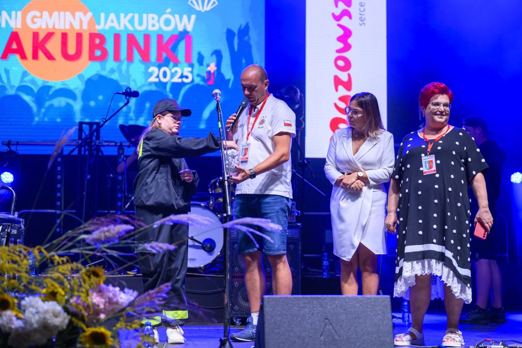 Na scenie cztery osoby, tło z niebieską grafiką. Kobieta w czerni podaje mikrofon mężczyźnie. Wokół jasne światła i kwiaty.