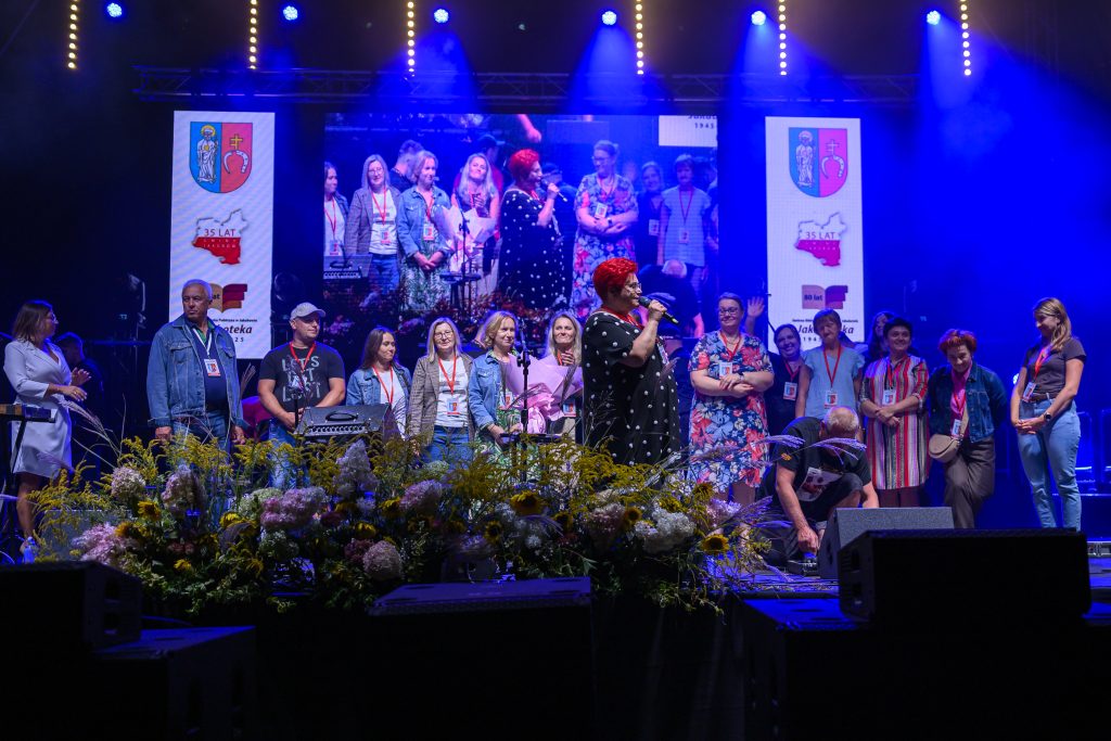 Osoby na scenie z mikrofonem, kolorowe światła i kwiaty z przodu. Ekran w tle pokazuje tę samą scenę. Rozświetlone tło w kolorze niebieskim.