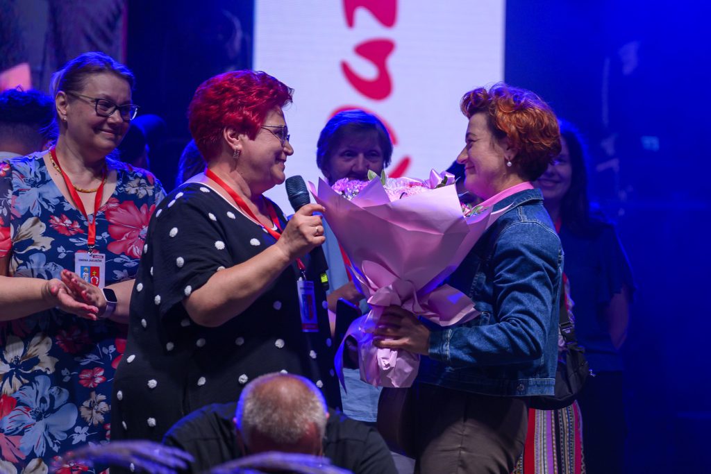 Dwie kobiety na scenie, z jednej trzymającej mikrofon, a druga bukiet kwiatów w różowym papierze. Tło w niebieskim świetle.