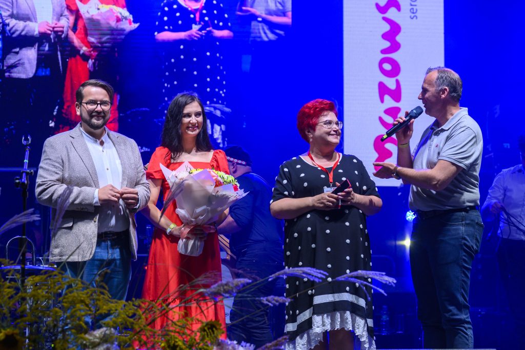 Na scenie stoją cztery osoby, jedna z nich trzyma mikrofon. W tle niebieskie światła i ekran z obrazem. Kobieta w czerwonej sukience trzyma kwiaty.