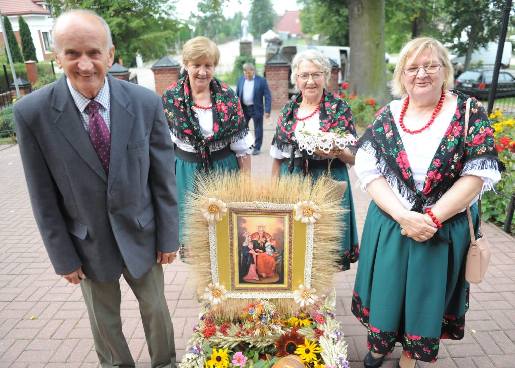 Na zdjęciu grupa ludzi w strojach ludowych z wieńcem zbożowym. Kolory są żywe; tło to zielony ogród. Centralnym elementem jest obraz w otoczeniu kwiatów.