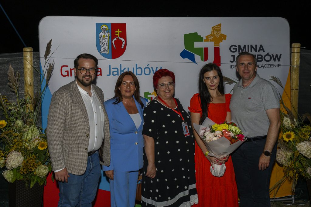 Grupa pięciu osób pozuje przed banerem Gmina Jakubów. Tło w jasnych barwach, osoby uśmiechnięte, jedna z bukietem.