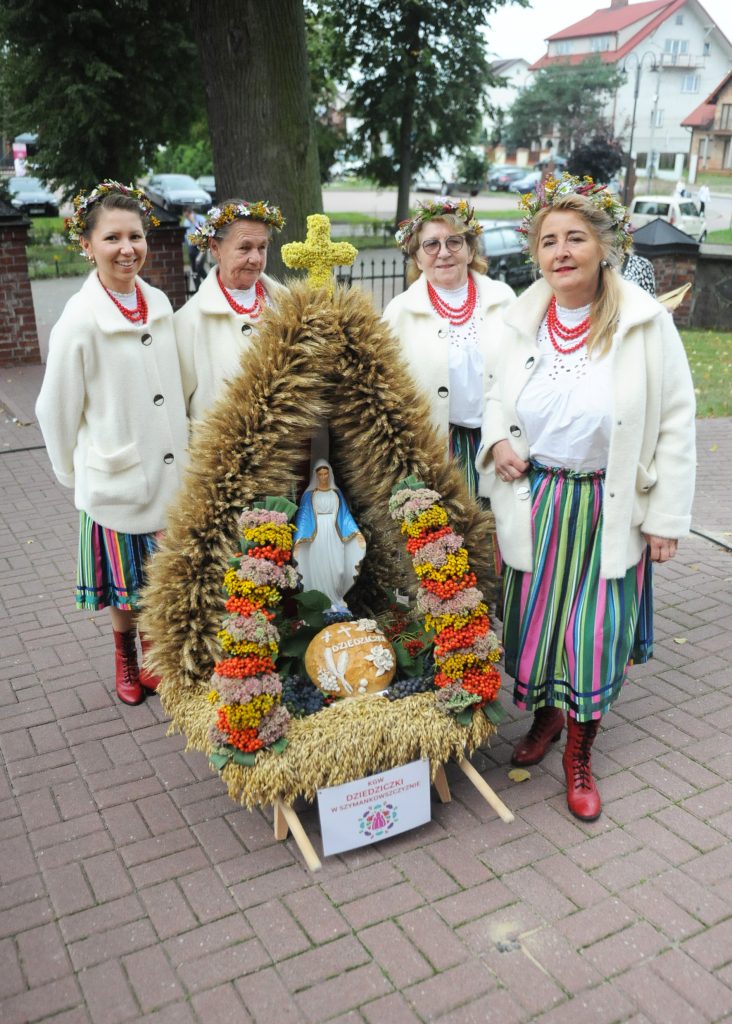 Cztery kobiety w tradycyjnych strojach stoją z wieńcem dożynkowym ozdobionym kwiatami i figurką Matki Boskiej. Stroje mają białe bluzki i kolorowe, pasiasta spódnice. Tło stanowi zielone drzewo i miejskie