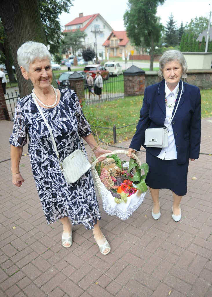 Dwie starsze kobiety niosą ozdobny kosz z kwiatami. Kobiety ubrane w eleganckie stroje, jedna w suknię, druga w garnitur. Tło z zielonymi drzewami i wiejskim domem.