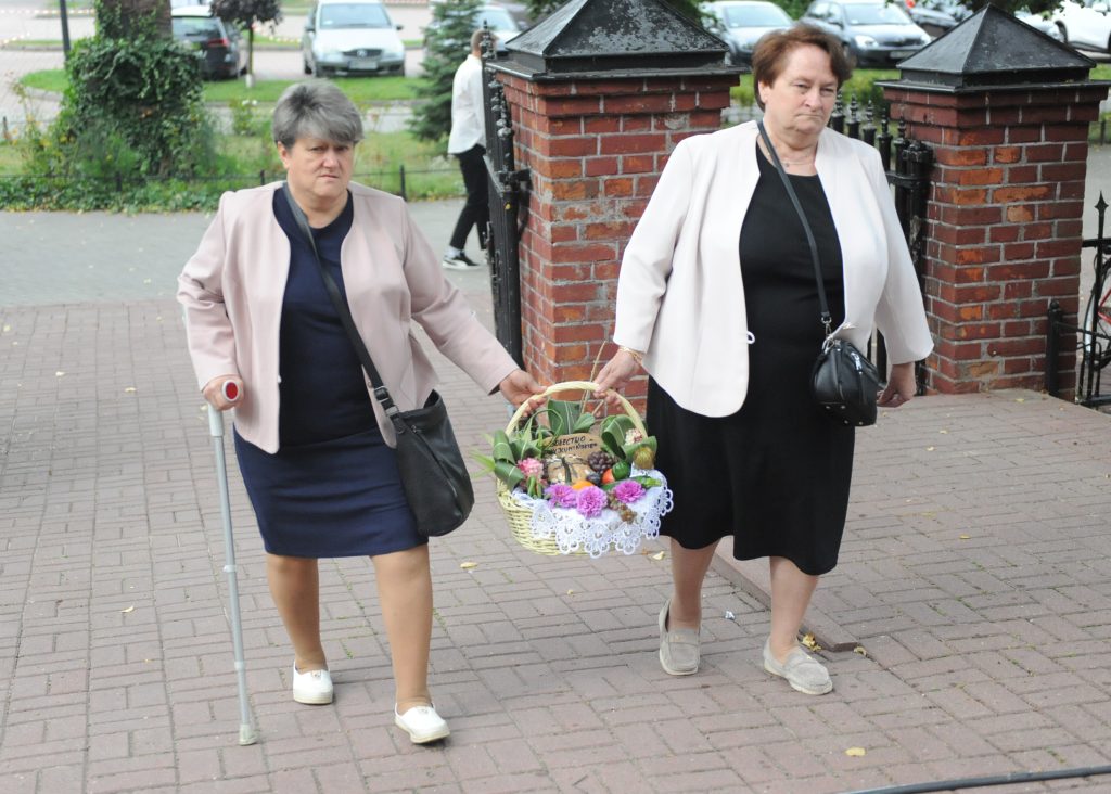 Dwie kobiety w jasnych żakietach idą chodnikiem z koszem kwiatów. Jedna z nich używa laski. W tle ceglana brama i zaparkowane auta.