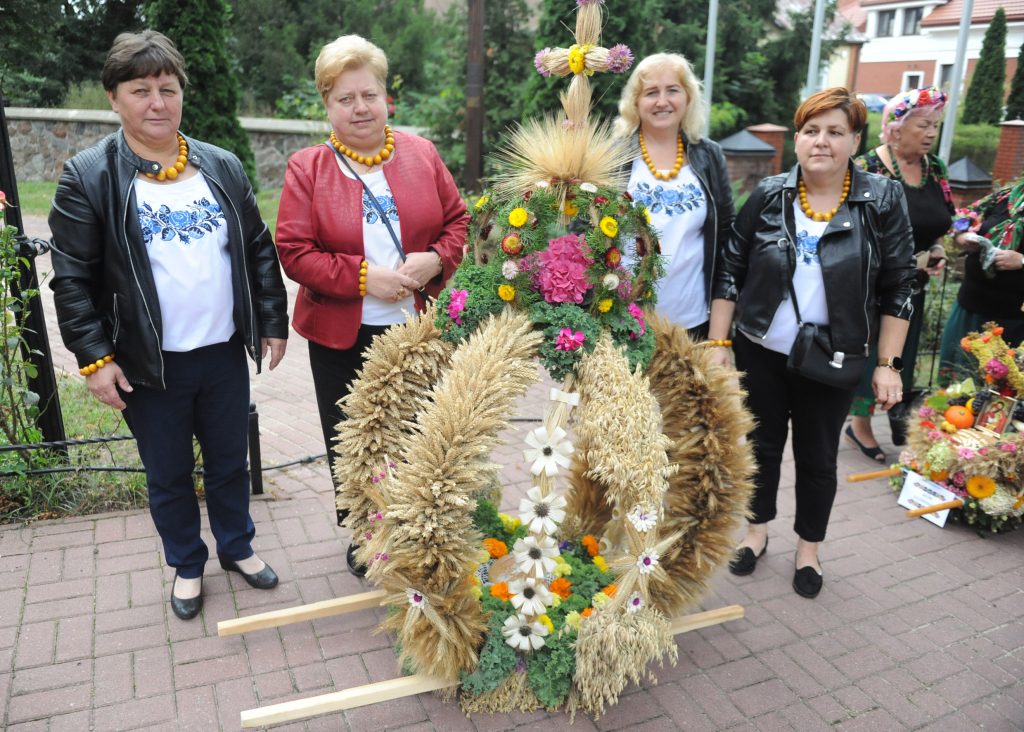 Panie w czarnych kurtkach stoją przy wieńcu dożynkowym zbożowo-kwiatowym. Tło jest zielone, widać krzewy i budynki. Kolory wyraziste, dominują złociste i zielone.