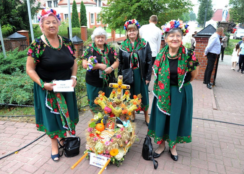 Cztery kobiety w ludowych strojach z kwiatowymi wiankami pozują z ozdobnym wózkiem. Tło z zielenią i ceglaną architekturą. Kolory: zielony, czerwony, wielobarwne kwiaty.