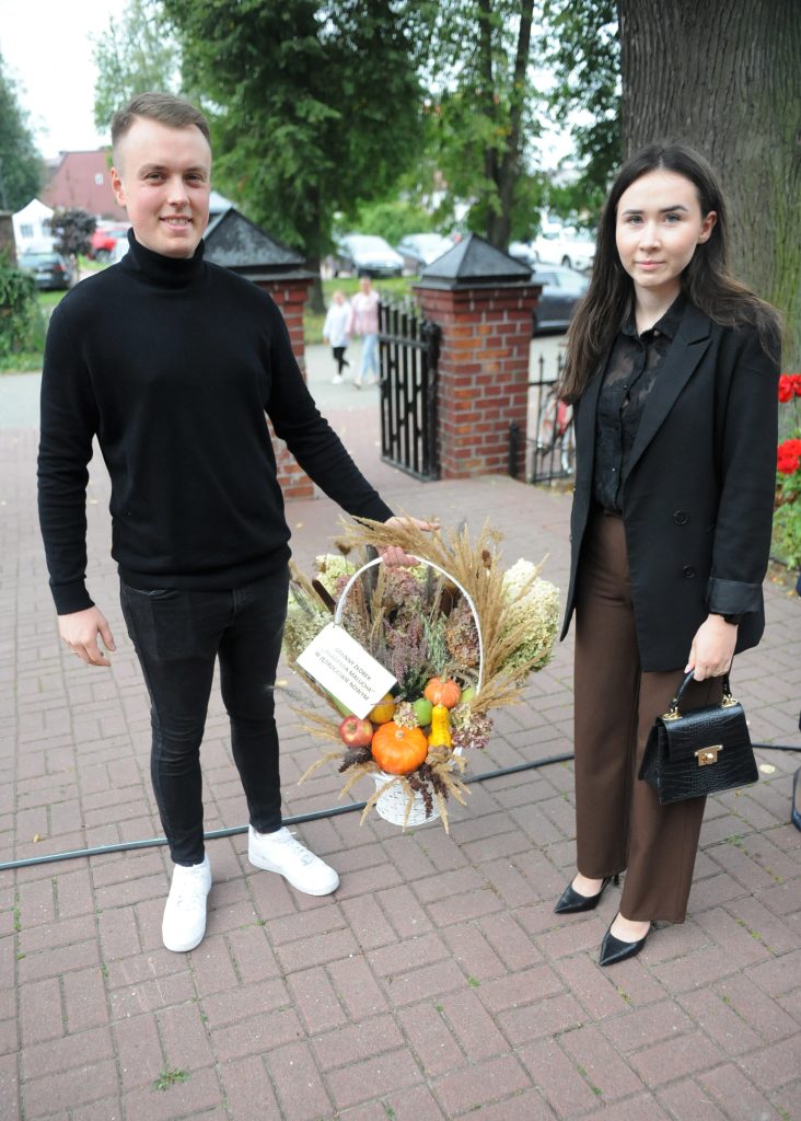 Para trzyma kosz z jesiennymi ozdobami na tle ceglanego muru i zieleni. Ubrani w ciemne kolory, stoją na bruku. Kosz zawiera dynie i suszone trawy.