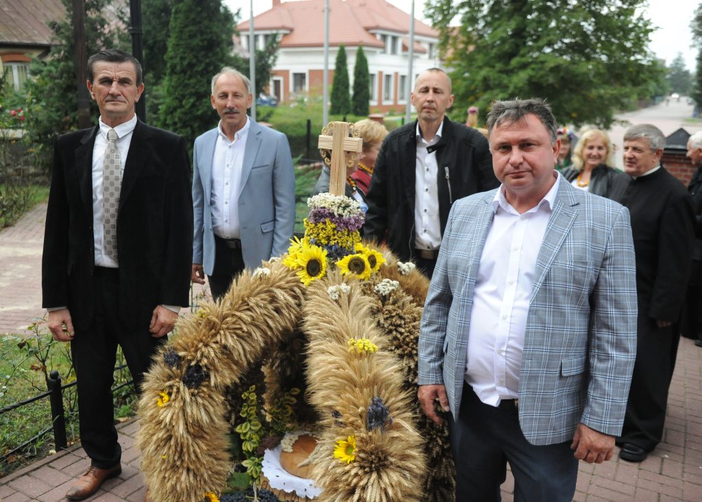 Na zdjęciu widać pięciu mężczyzn w garniturach. Jeden trzyma dożynkową dekorację z pszenicą i słonecznikami. W tle są zielone drzewa i domy.