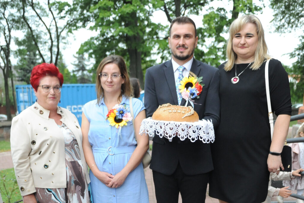 Czworo osób stoi na zewnątrz przed drzewami. Mężczyzna trzyma chleb na serwecie. Kobiety są ubrane w różne sukienki i marynarki.