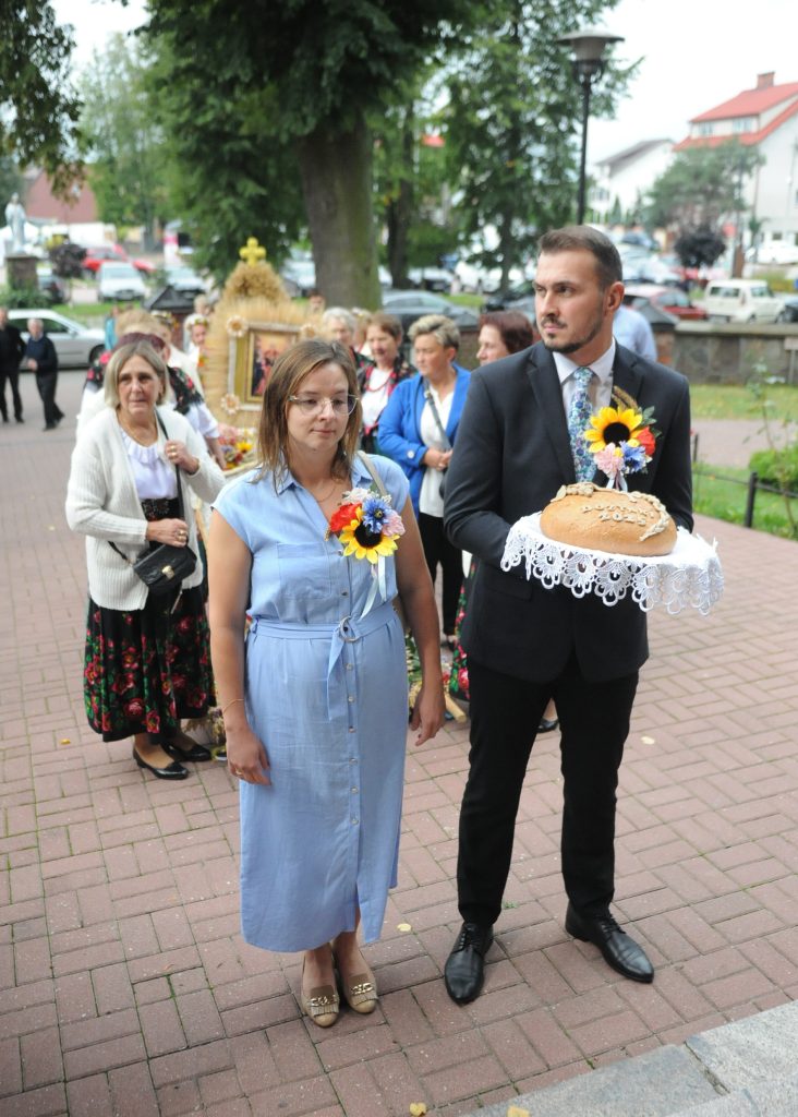 Na zdjęciu widnieje para z chlebem ozdobionym kwiatem. Kobieta ma niebieską sukienkę, mężczyzna czarny garnitur. W tle jest grupa osób, drzewa i budynki.
