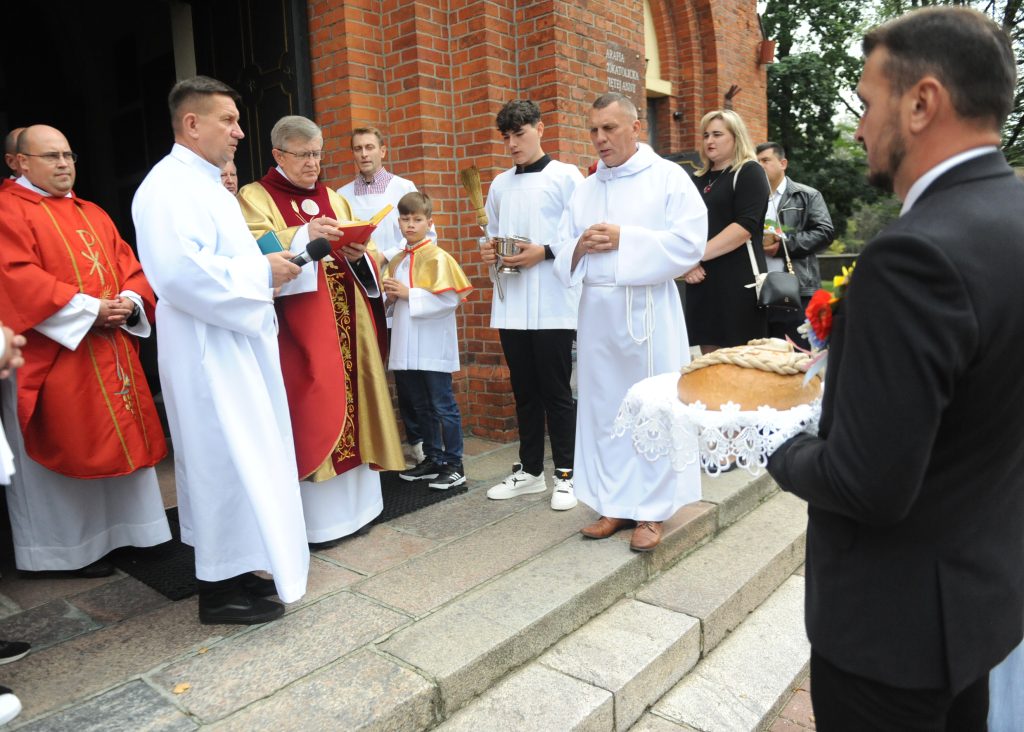Grupa osób w białych i czerwonych szatach stoi przed ceglaną budowlą. W tle zielone drzewo. Mężczyzna trzyma chleb na tacy.