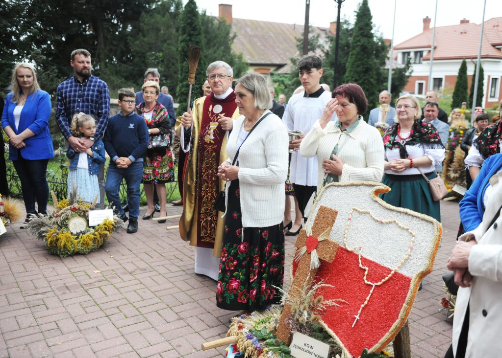 Grupa ludzi uczestniczy w religijnej uroczystości na zewnątrz. Widoczne barwne stroje ludowe i wieńce żniwne. Tło z drzewami i budynkami.