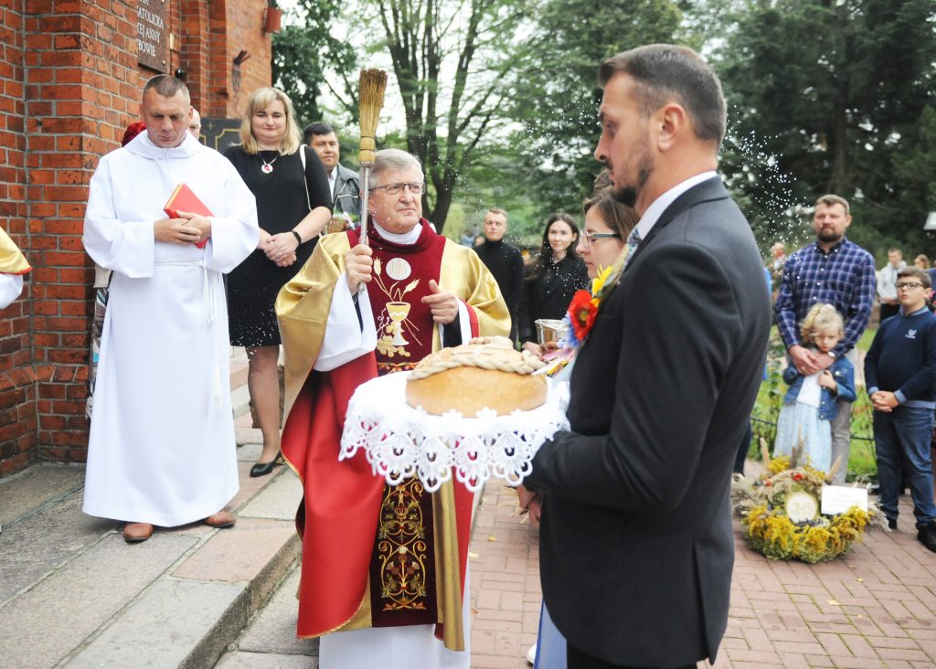 Osoby przed kościołem podczas ceremonii. Ksiądz w szatach liturgicznych święci chleb. W tle drzewa i ludzie.