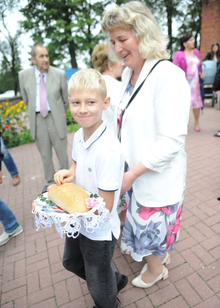Chłopiec w białej koszuli trzyma chleb na koronkowej serwetce. Obok stoi kobieta w białej marynarce i kolorowej sukience. Tło to brukowany chodnik z kwiatami i ludźmi.