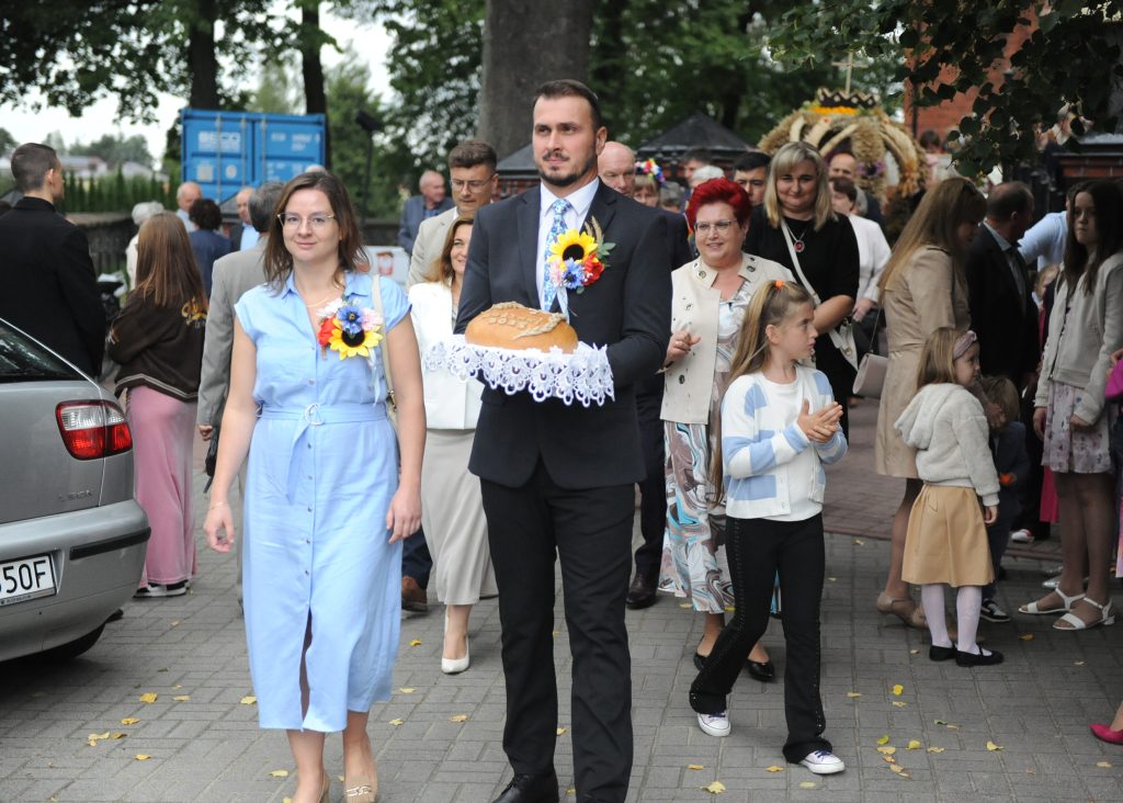 Na zdjęciu widać ludzi idących w procesji z chlebem na tacy. Mężczyzna w garniturze i kobieta w jasnej sukience prowadzą grupę. Tło to zielone drzewa i tłum uczestników.