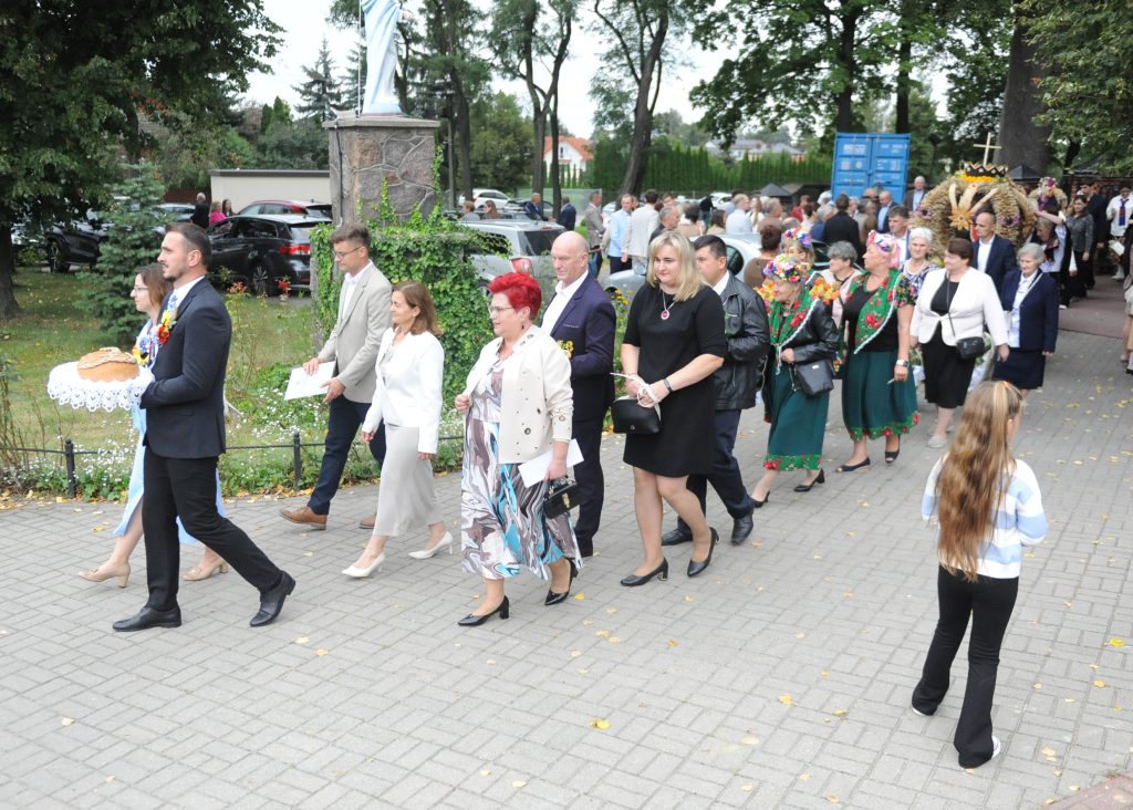 Grupa ludzi na spacerze w parku. Ciemne i jasne ubrania, zielone tło z drzewami. Niesiony chleb na białej serwetce.