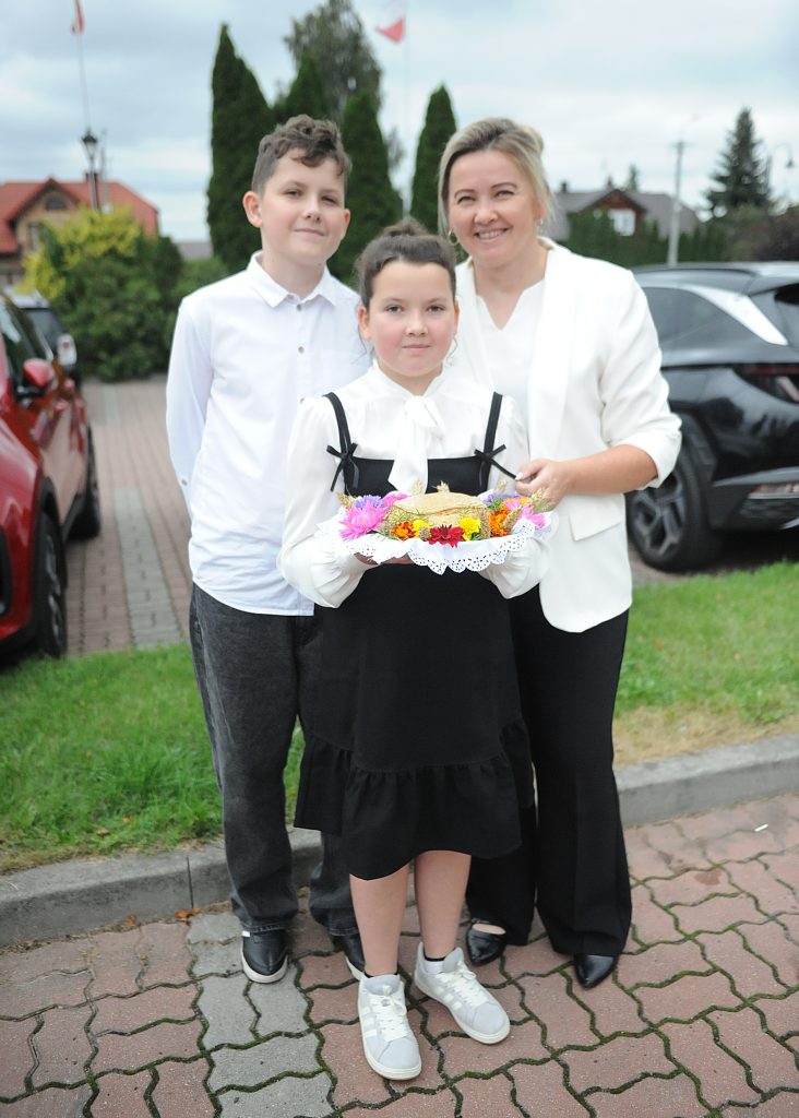 Trzy osoby w eleganckiej odzieży stoją na zewnątrz. Dziewczynka trzyma kolorowy wieniec z kwiatami. W tle są samochody i drzewa.