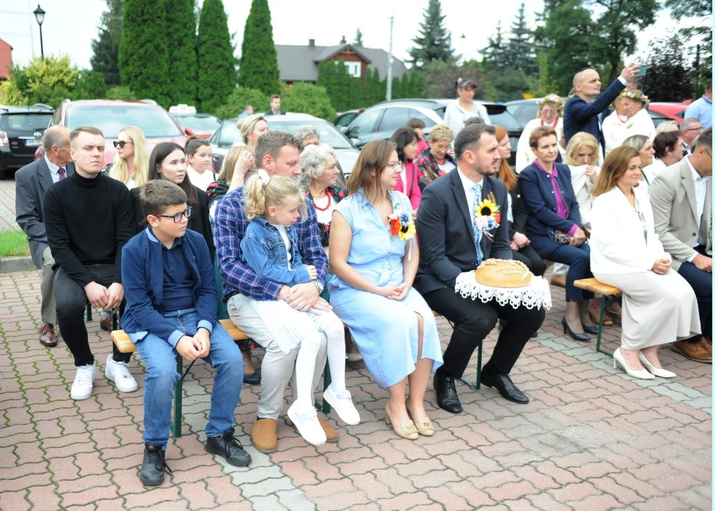 Grupa ludzi siedzi na ławkach podczas wydarzenia na świeżym powietrzu. Mężczyzna trzyma chleb, tło to zaparkowane auta i krzewy. Kolory są jasne, dominują błękit i zieleń.