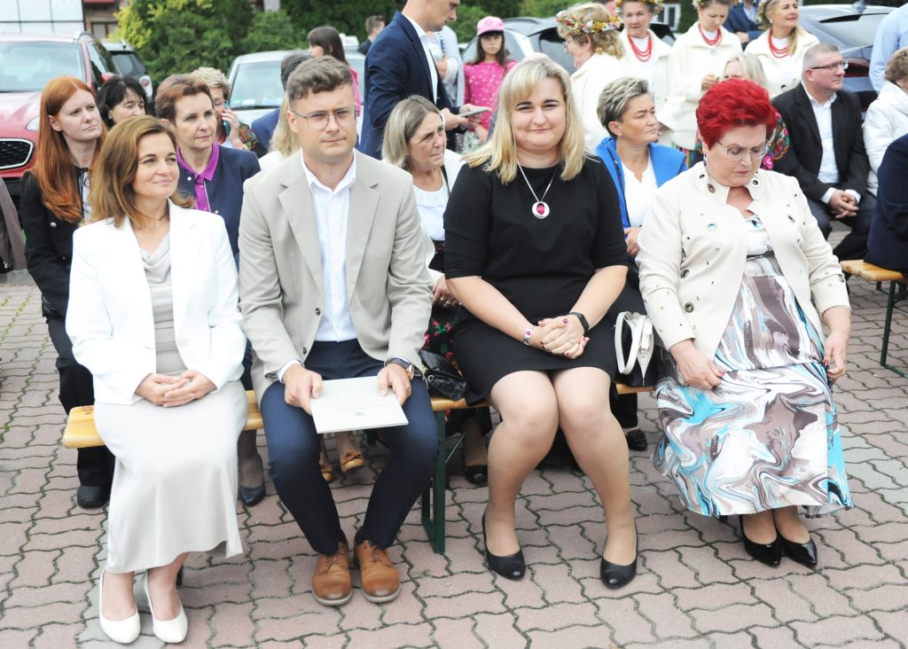 Grupa osób siedzi na ławkach na zewnątrz. Są ubrane elegancko, a w tle widać samochody i zieleń. Kobiety mają na sobie jasne stylizacje.