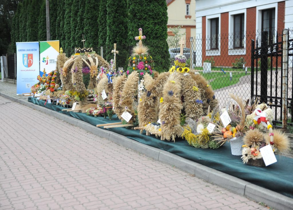 Świąteczne wieńce zbożowe z kwiatami na chodniku, obok zielone drzewa i banery. Tło stanowią ogrodzenie i budynek z cegły. Kolory to głównie brązy, zielenie i żółcie.