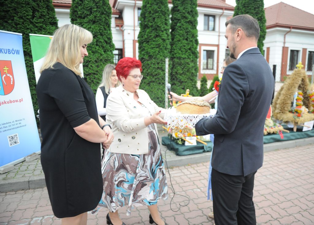Grupa osób stoi na zewnątrz przy straganach, jedna z nich trzyma bochenek chleba na białej serwetce. Za nimi zielone tuje i biały budynek z czerwonym dachem. Kobieta w jasnej marynarce wręcza chleb mężczyźnie