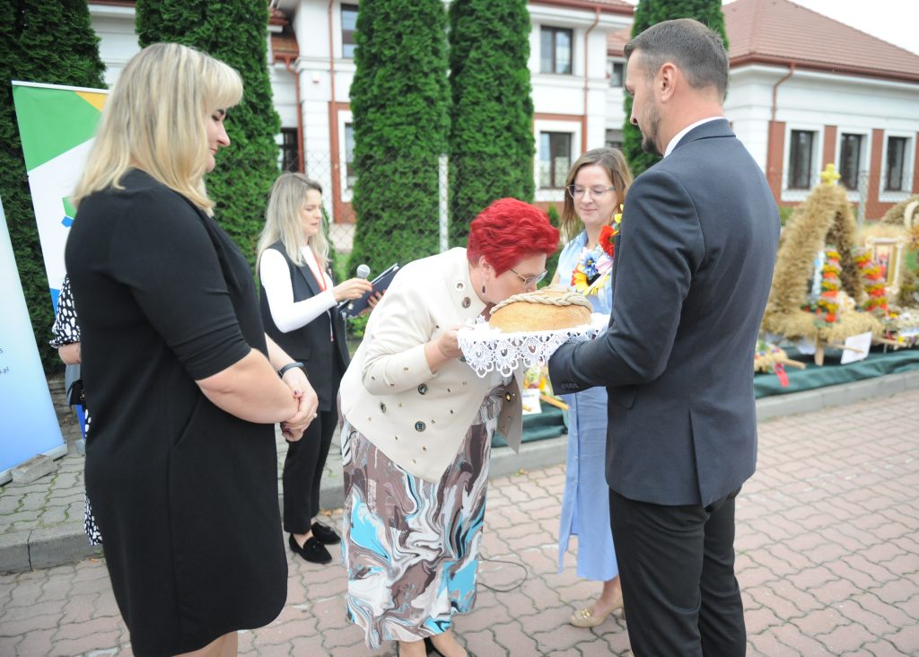 Na zdjęciu grupa osób uczestniczy w ceremonii na zewnątrz. Kobieta z czerwonymi włosami całuje chleb na białej serwecie. Tło tworzą zielone drzewa i budynek.