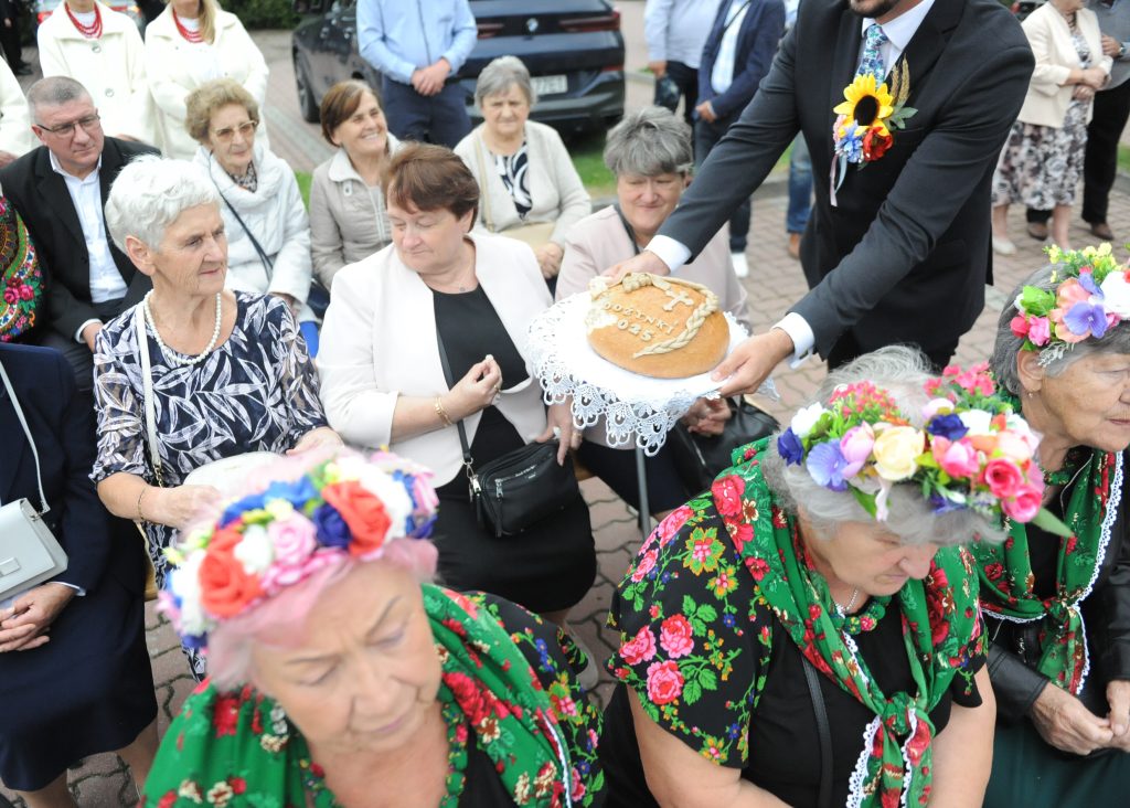 Na zdjęciu grupa starszych osób, w tym kobiety w kolorowych wiankach i chustach. Mężczyzna w garniturze podaje chleb z napisem Dożynki 2023. Tło stanowią ludzie na tle parku i samochodu.