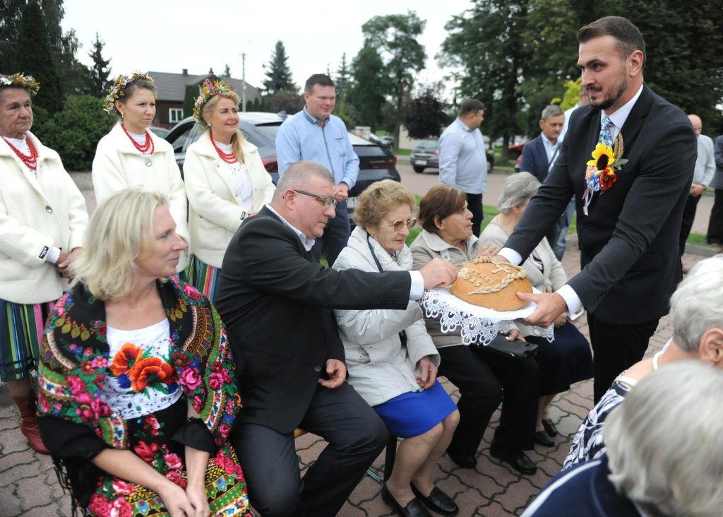 Grupa ludzi w tradycyjnych strojach na zewnątrz. Mężczyzna podaje chleb na tacy. Tło z drzewami i samochodami.