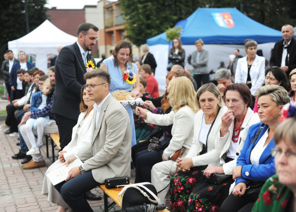 Na zdjęciu widać grupę ludzi siedzących na ławkach podczas plenerowego wydarzenia. Osoby są ubrane elegancko, przeważają jasne kolory ubrań. W tle widać niebieskie i białe namioty.